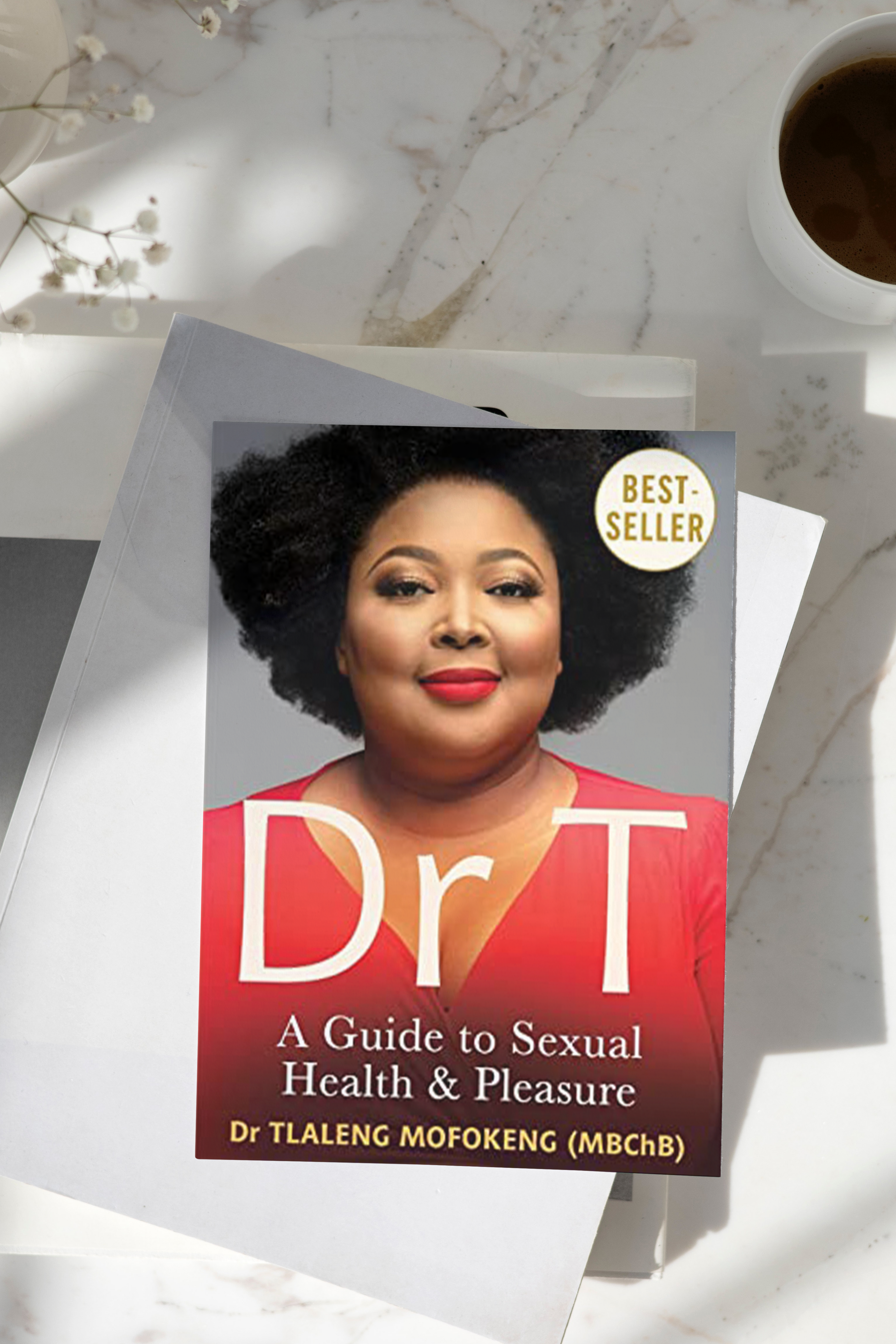 A book titled "Dr T: A Guide to Sexual Health & Pleasure" by Dr. Tlaleng Mofokeng is displayed on a white marble surface next to a cup of coffee and small white flowers.